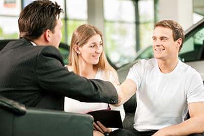 Dealership, the dealer is handing auto keys to a young couple, they are excited, cars standing in the background